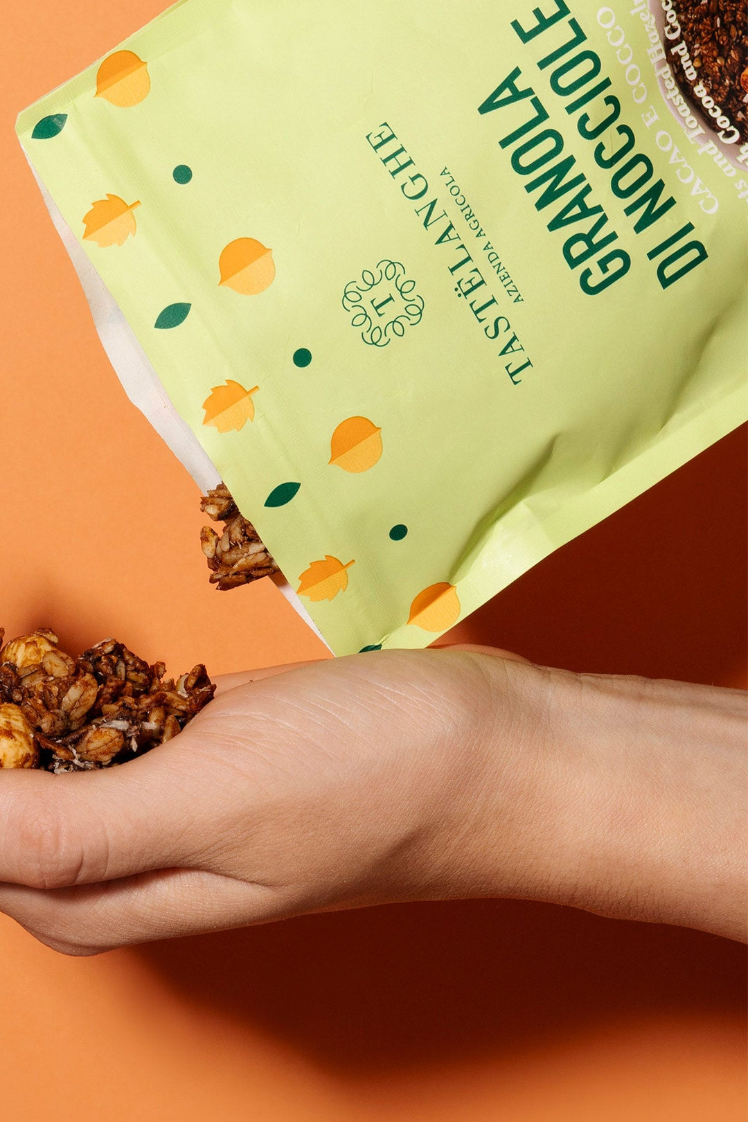 Granola di nocciole Tastëlanghe cacao e cocco versata dalla confezione verde in una mano, su fondo arancio.