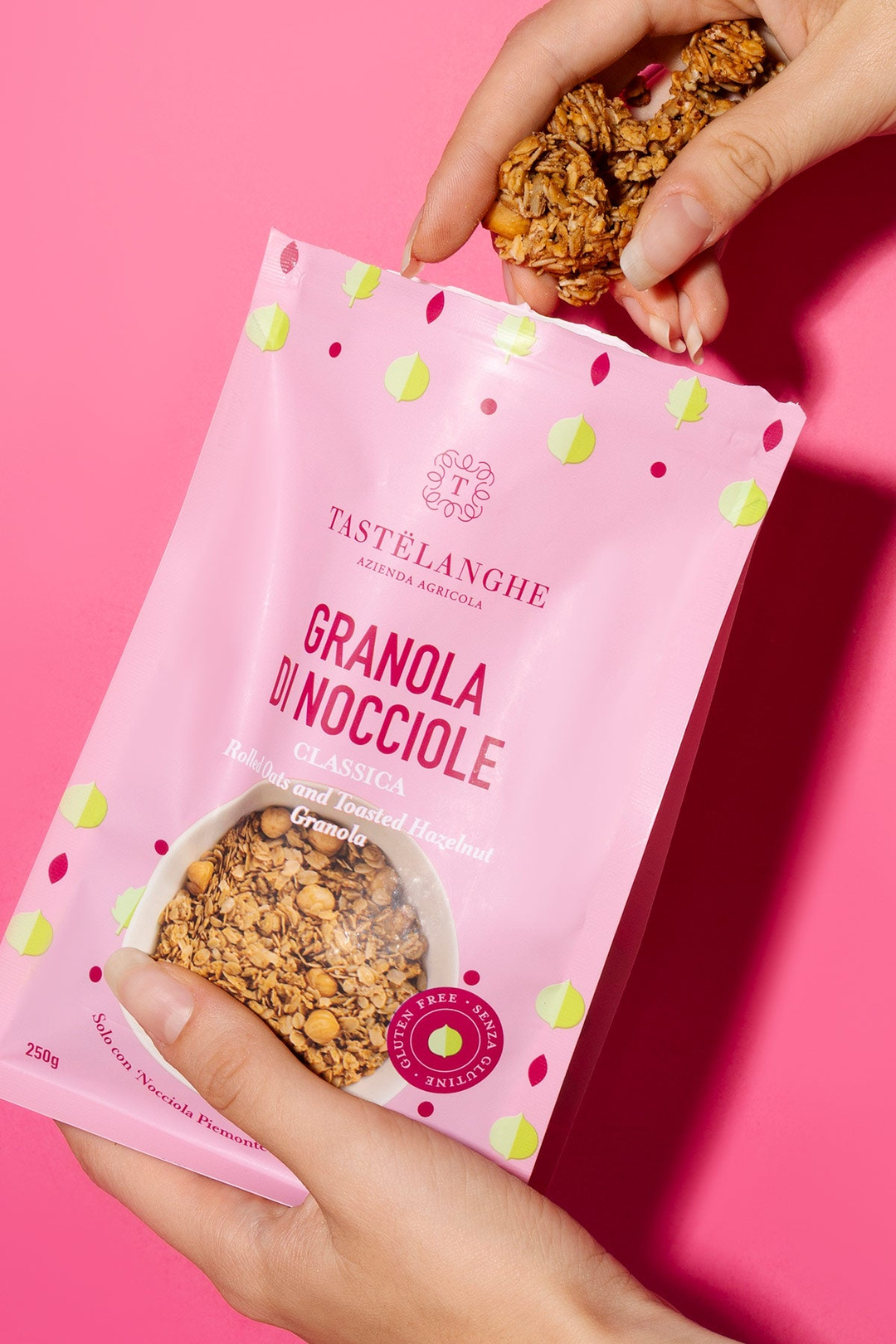 Due mani presentano la confezione rosa di granola classica Tastëlanghe e una porzione di granola, su fondo rosa.