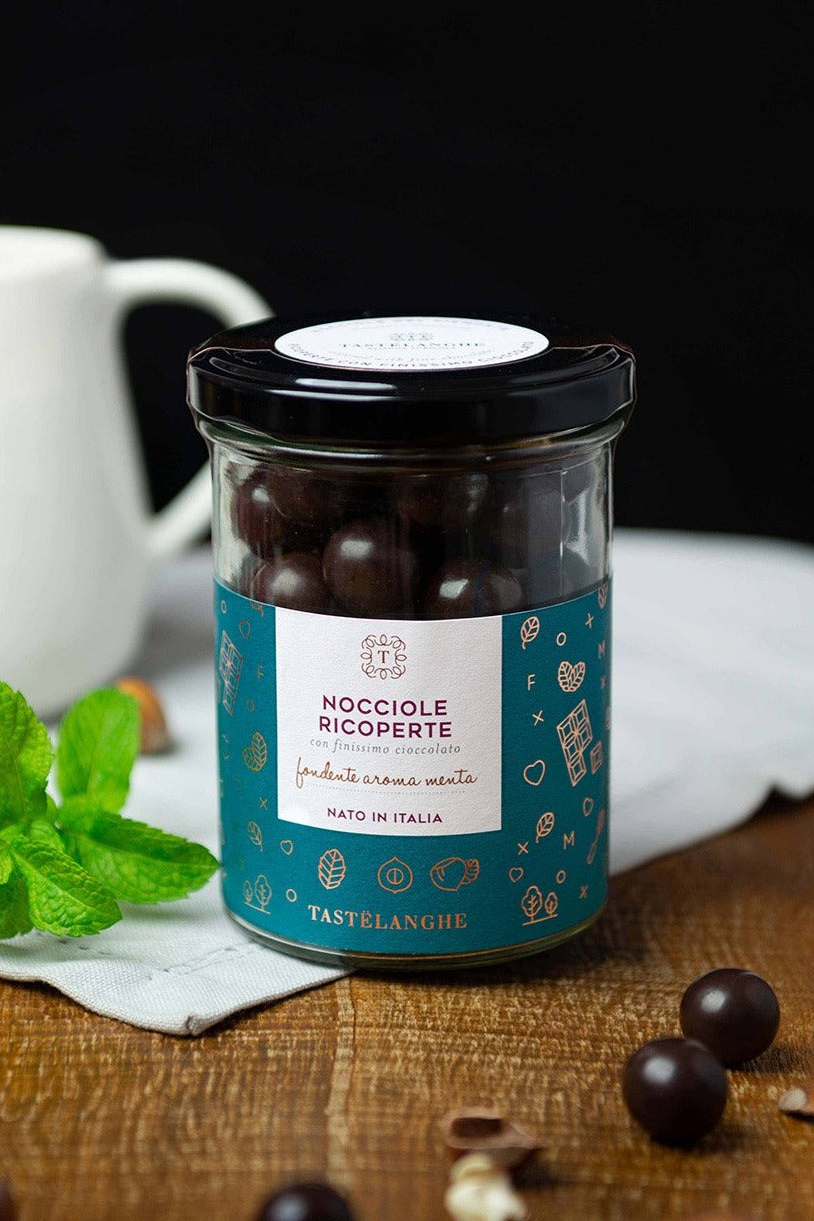 Vasetto Tastëlanghe di nocciole ricoperte di cioccolato fondente alla menta, con foglie di menta su un tavolo in legno.