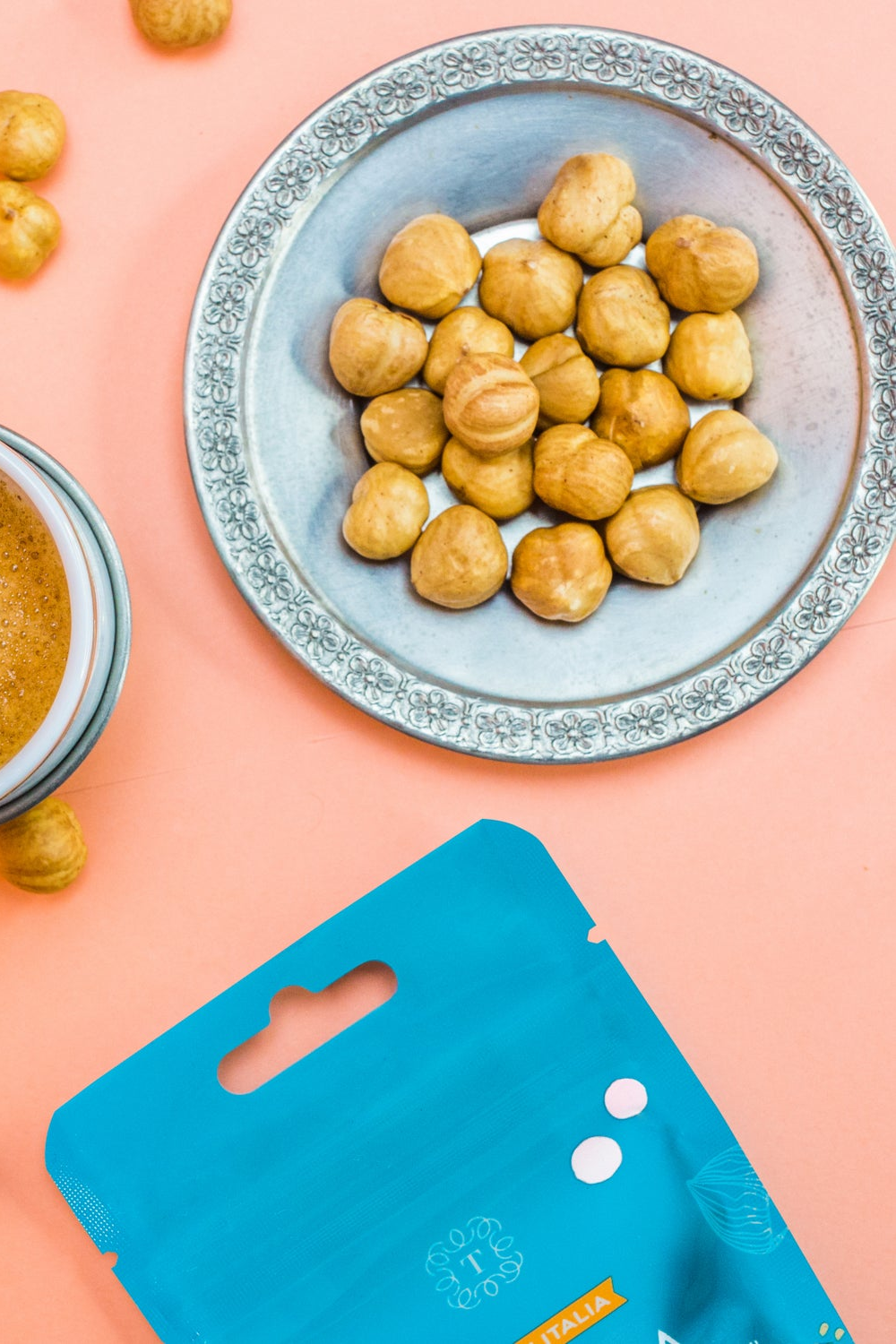 Nocciole tostate su un piatto argentato, una tazza di caffè e una confezione snack Tastëlanghe su fondo pesca.