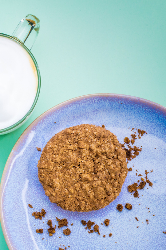 Tortino di nocciole su un piatto lilla con briciole, accanto a un cappuccino su fondo verde acqua.